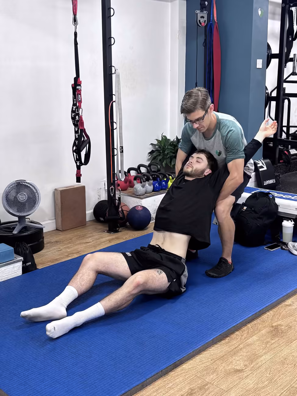 Physiotherapist supporting a client’s upper back and arm through an assisted trunk stretch on a mat in the Exeter studio