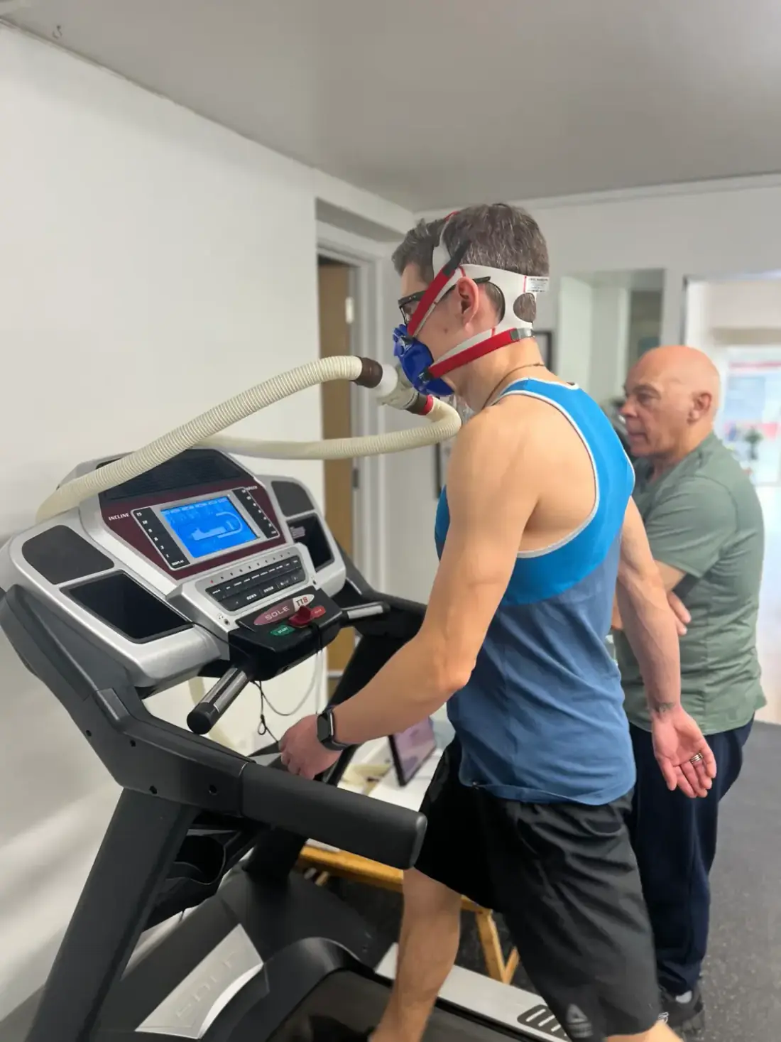 Man walking on a treadmill wearing a breath-analysis mask and tube while the tester stands beside him monitoring the VO2 max test