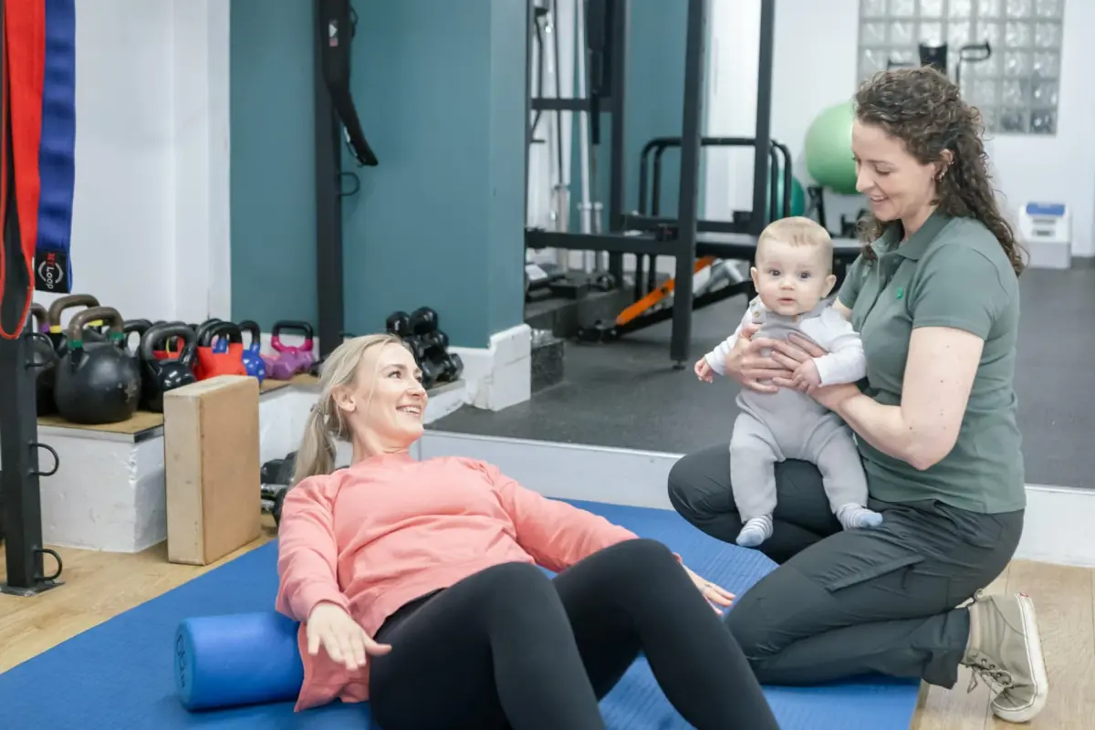 Women’s health physiotherapist holding a smiling baby while the baby’s mother lies back on a foam roller during a postnatal exercise session